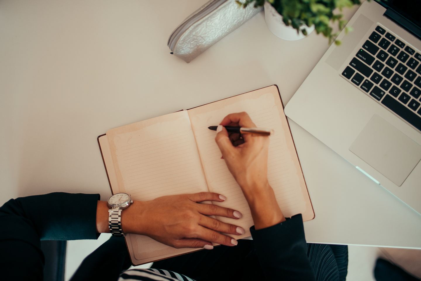 Manos escribiendo en un cuaderno abierto junto a un portátil