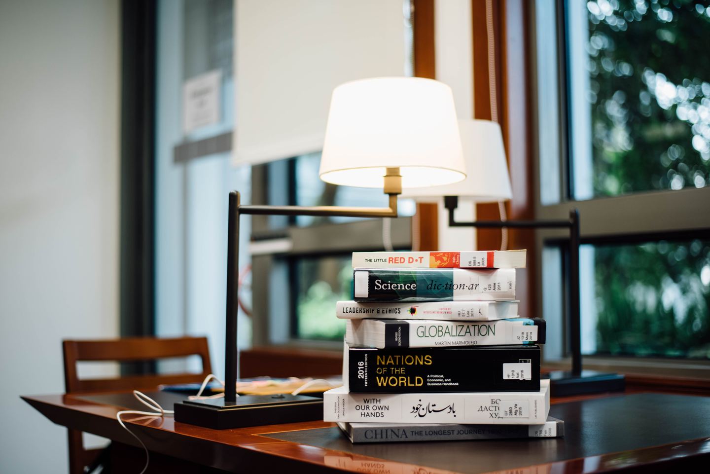 Libros apilados junto a una lámpara encendida en una mesa frente a la ventana