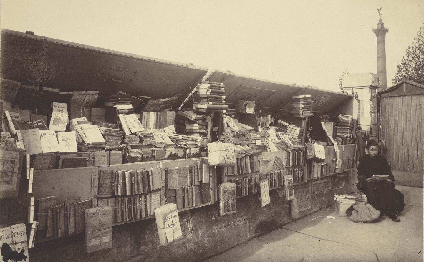 Puesto de libros viejos al aire libre con los cajones abiertos y llenos de volúmenes, una mujer sentada leyendo junto a ellos y la columna de la Bastilla al fondo