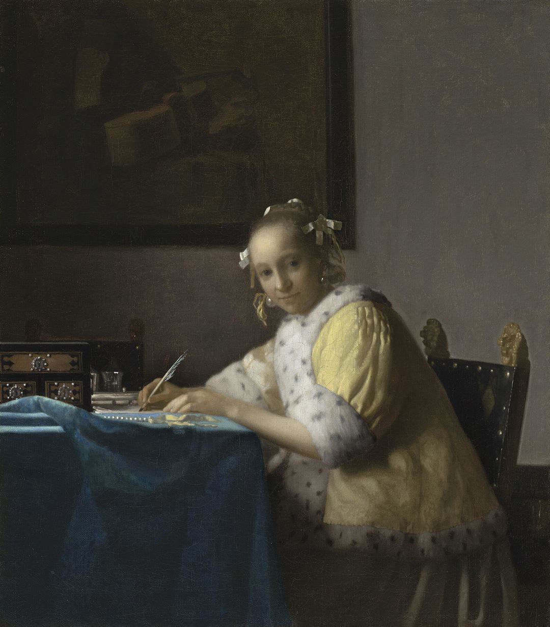 Mujer con chaqueta amarilla escribiendo una carta en una mesa y mirando hacia el espectador